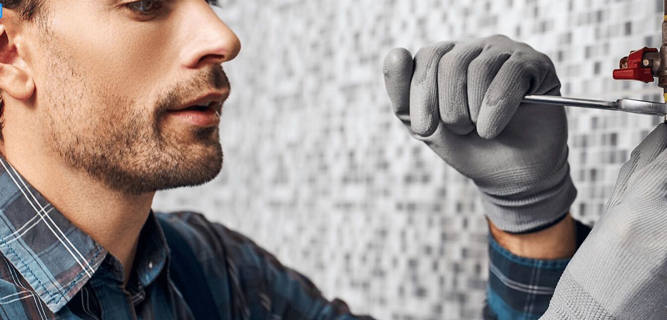Handwerker mit grauen Handschuhen arbeitet mit Schraubendreher an Wasserleitung vor gefliester Wand